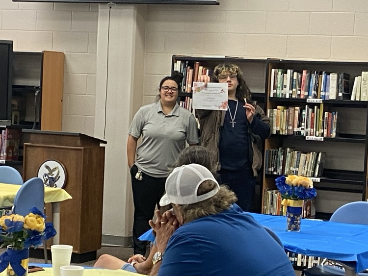 a student in black pants, a blue shirt, and brown jacket is presented a certificate by Ms. McMillian in black pants and a grey shirt.