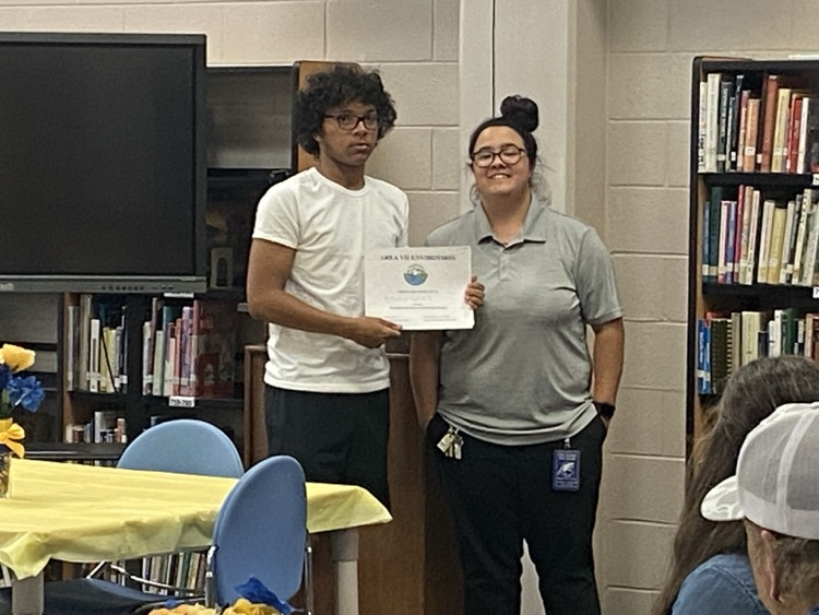a student in a black shorts and a white tshirt is presented a certificate by Ms. McMillian in black pants and a grey shirt.