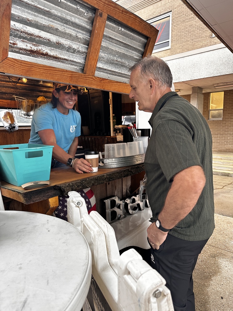 A barista in a blue t-shirt smiles from behind the counter of the Brew mobile coffee trailer while serving a staff member.