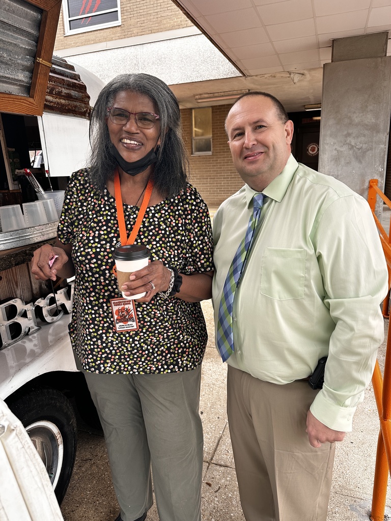 Two staff members smile together outside near the Brew mobile coffee trailer. One holds a hot coffee cup and wears a Cougars lanyard.