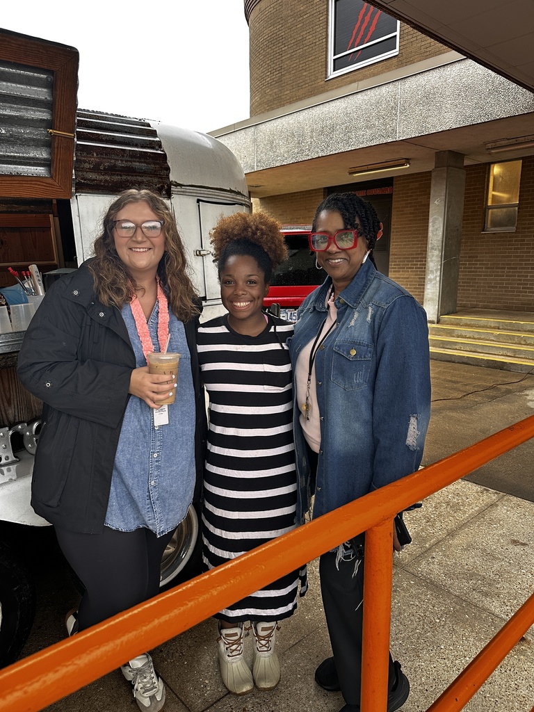 Three staff members smile together outside near the Brew coffee trailer. One holds an iced coffee drink.