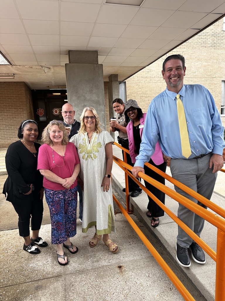 A group of seven staff members poses on the school's front steps and ramp area, smiling together.