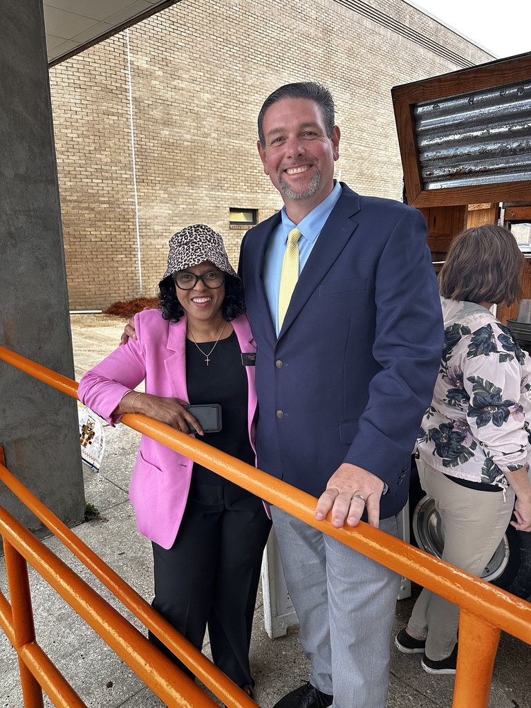 Two staff members pose together beside the orange railing outside the school, smiling. A woman in a pink blazer and leopard-print hat stands next to a man in a navy suit with a yellow tie.