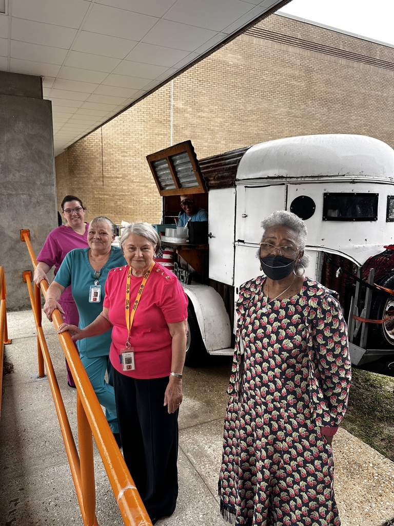 Four staff members line up along the orange railing outside the school near the Brew coffee trailer, smiling for the photo.