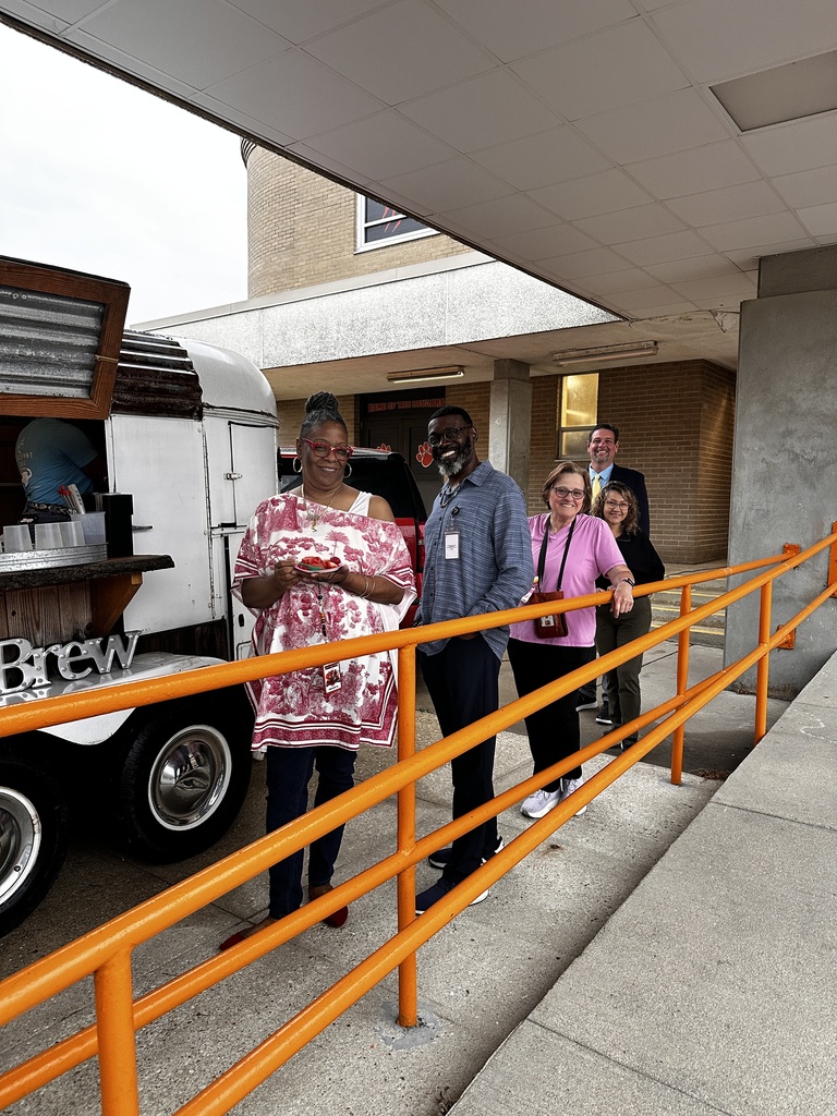 A group of four staff members smile together outside a school building near a mobile coffee trailer called "Brew." One person holds a small plate of food.