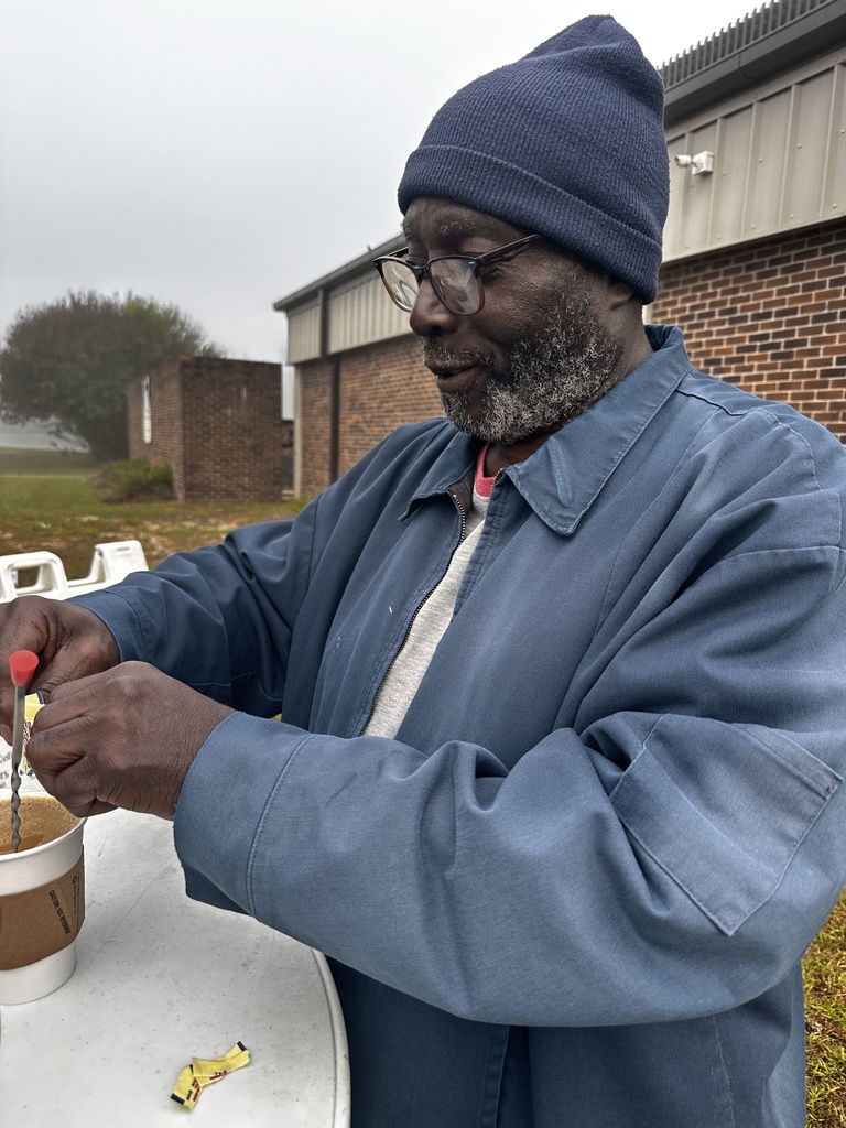 A staff member in a blue jacket and knit cap stirs sugar into his coffee at an outdoor table on a cool, foggy morning.