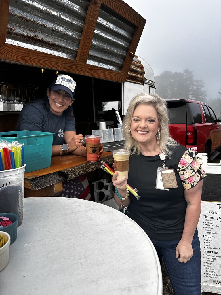 A smiling staff member holds an iced coffee drink at the Brew mobile coffee trailer window while the barista smiles from inside. The trailer's menu listing hot coffee, frappes, and smoothies is visible nearby.