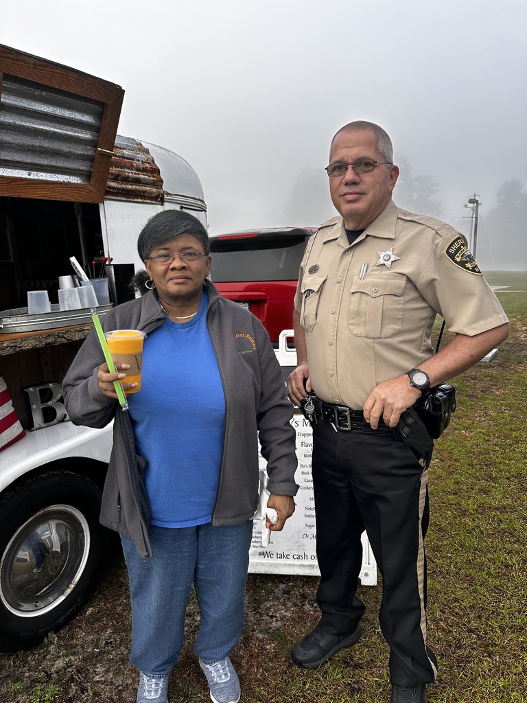 A staff member holding an iced drink poses with a sheriff's deputy beside the Brew mobile coffee trailer on a foggy morning. The trailer's menu board is visible in the background.