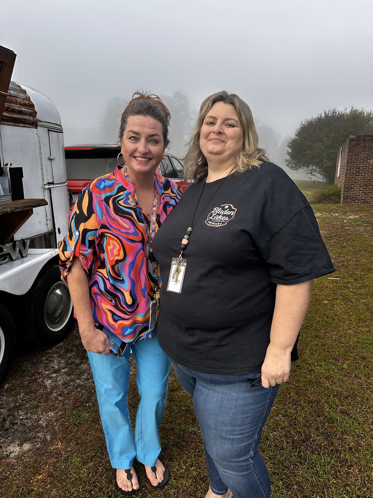 Two staff members smile together outdoors on a foggy morning near the Brew mobile coffee trailer. One wears a colorful swirl-print top and turquoise pants; the other wears a black Bladen Lakes Primary shirt.