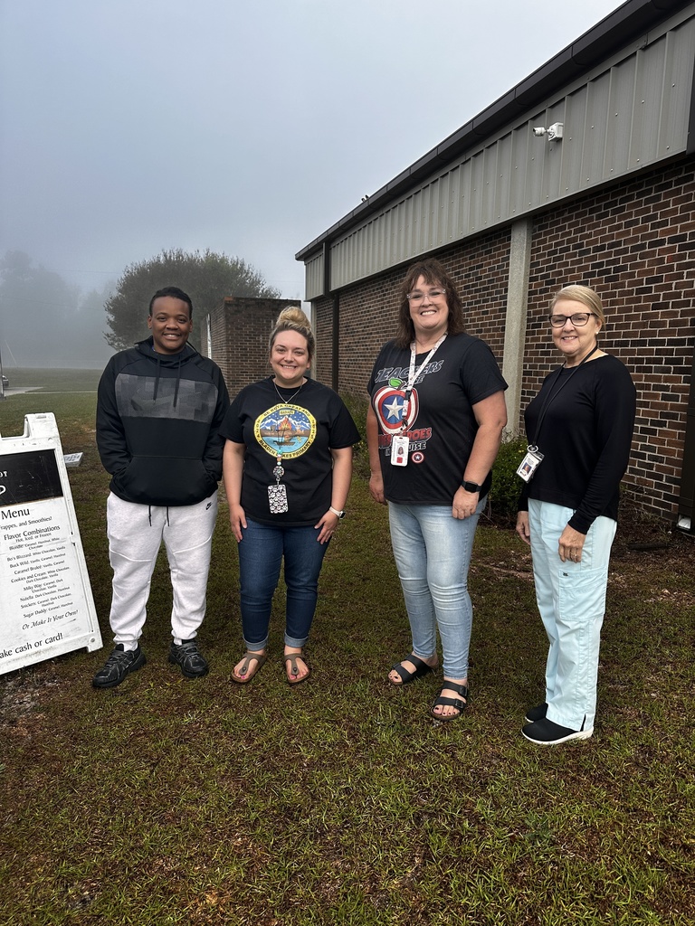 Four staff members smile together outside a brick school building on a foggy morning, standing near the Brew coffee trailer's menu board. Two wear Bladen Lakes Primary shirts.