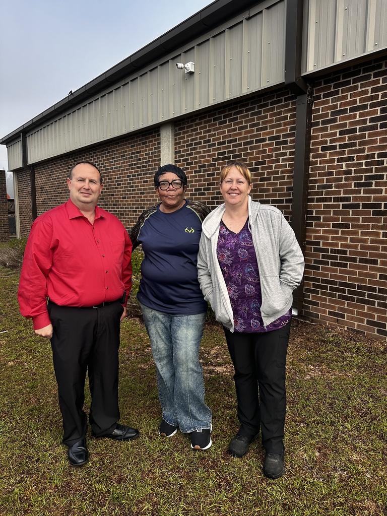  Three staff members pose together outside a brick school building on a foggy morning, smiling at the camera.