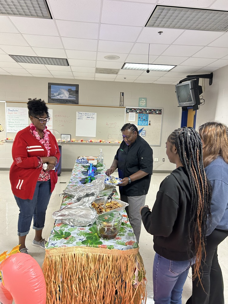 aloha themed table with grass skirt around it. two pans of food as a chef serves a teacher. two students stand nearby to help serve! 