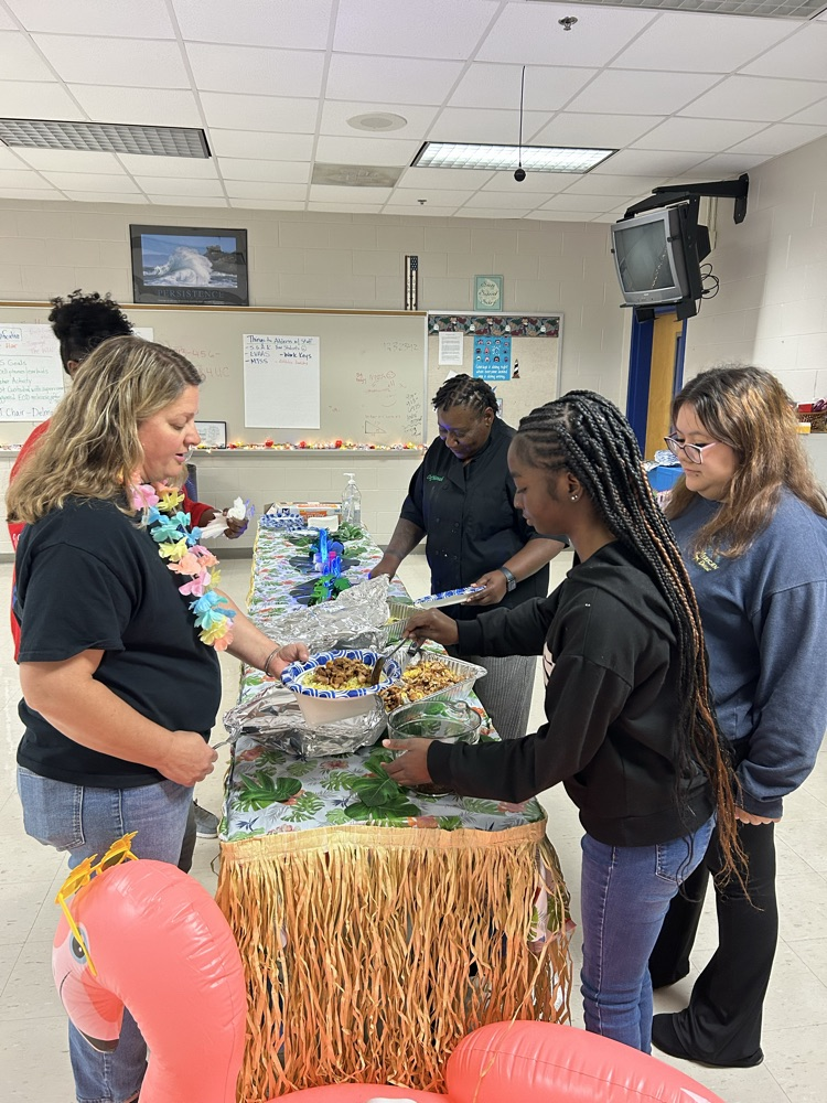 aloha themed table with grass skirt around it. two pans of food as a chef serves a teacher. two students stand nearby to help serve! 