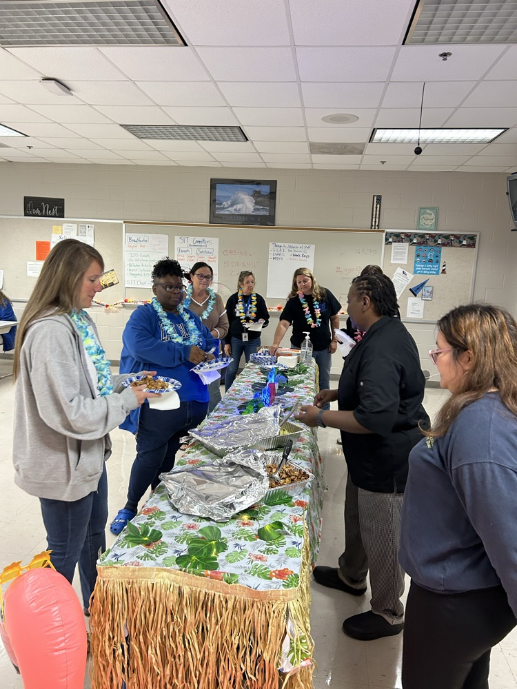 aloha themed table with grass skirt around it. two pans of food as a chef serves a teacher. two students stand nearby to help serve! 