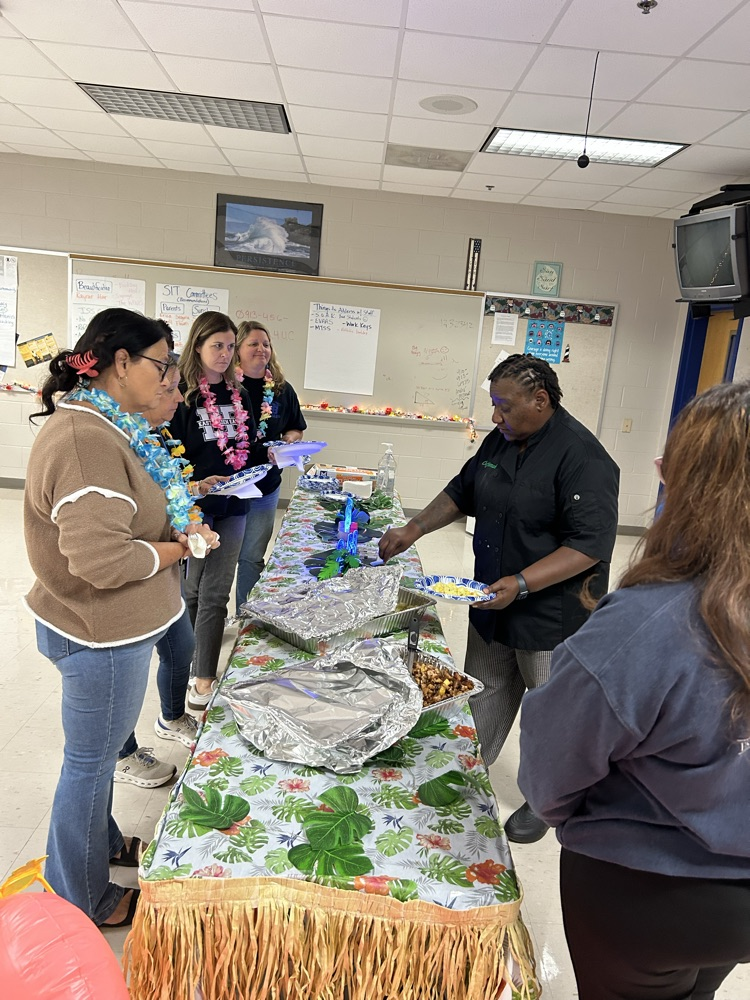 aloha themed table with grass skirt around it. two pans of food as a chef serves a teacher. two students stand nearby to help serve! 