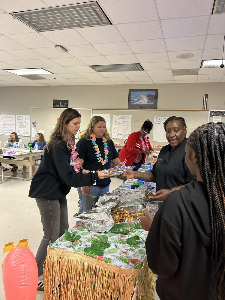 aloha themed table with grass skirt around it. two pans of food as a chef serves a teacher. two students stand nearby to help serve! 