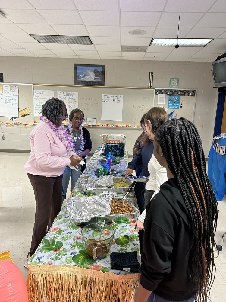 aloha themed table with grass skirt around it. two pans of food as a chef serves a teacher. two students stand nearby to help serve! 