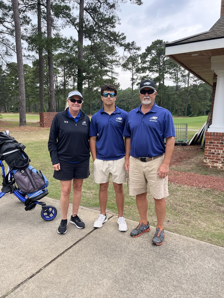 Coach Patty Evers, student athlete Henry Simmons, and golf Coach Huff standing together on the gold course as they stand beside a blue golf bag  