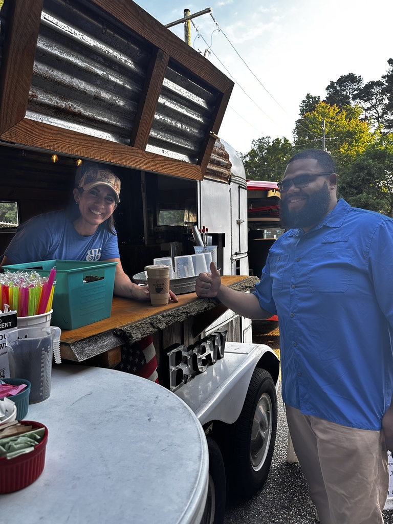 A woman working inside a mobile coffee trailer called Barefoot Brew smiles and leans on the wooden counter while a man in a blue button-down shirt gives a thumbs up. Colorful straws and drink supplies are visible on the counter.