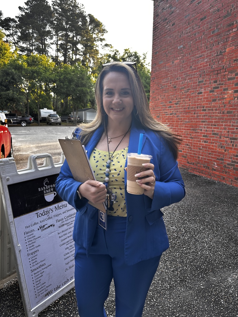 A woman in a royal blue blazer and yellow patterned top smiles while holding an iced coffee drink and a clipboard, standing next to a Barefoot Brew mobile coffee trailer menu sign.