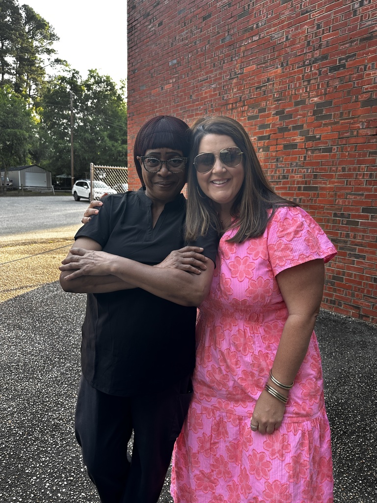 Two women stand with arms around each other smiling in front of a red brick wall. One wears an all-black outfit and the other wears a bright pink floral dress with gold bangle bracelets.