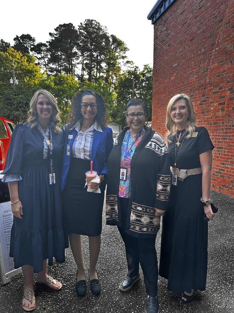 Four women wearing lanyards and ID badges smile together outdoors next to a brick building, with a Barefoot Brew menu sign partially visible at left.