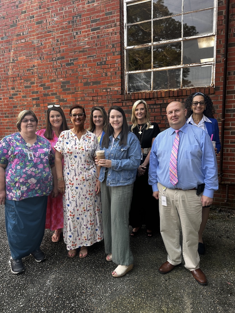 A group of eight school staff members — seven women and one man — pose together smiling in front of a red brick building. Several are holding iced coffee drinks and wearing ID lanyards.