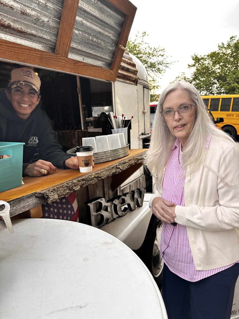 A woman with long white hair and glasses stands at the window of the Barefoot Brew coffee trailer, where a smiling barista in a cap hands her a branded cup. A North Carolina school bus is visible in the background.