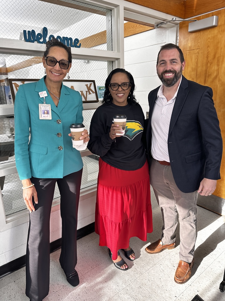 Three people — a woman in a teal blazer, a woman in a black Seahawks sweatshirt and red skirt holding a Barefoot Brew coffee cup, and a man in a navy blazer — pose together smiling inside a school entrance with a "Welcome" sign visible behind them.