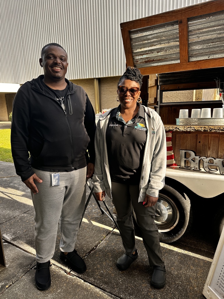 A man in a black hoodie and a woman wearing a "DeAnna" name tag and Nighthawks Housing Coalition polo stand together smiling in front of the Barefoot Brew coffee trailer.