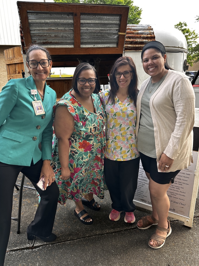 Four women pose together smiling in front of the Barefoot Brew coffee trailer in a school parking lot. They are dressed in colorful attire, including a teal blazer and floral prints. A menu board is partially visible to the right.