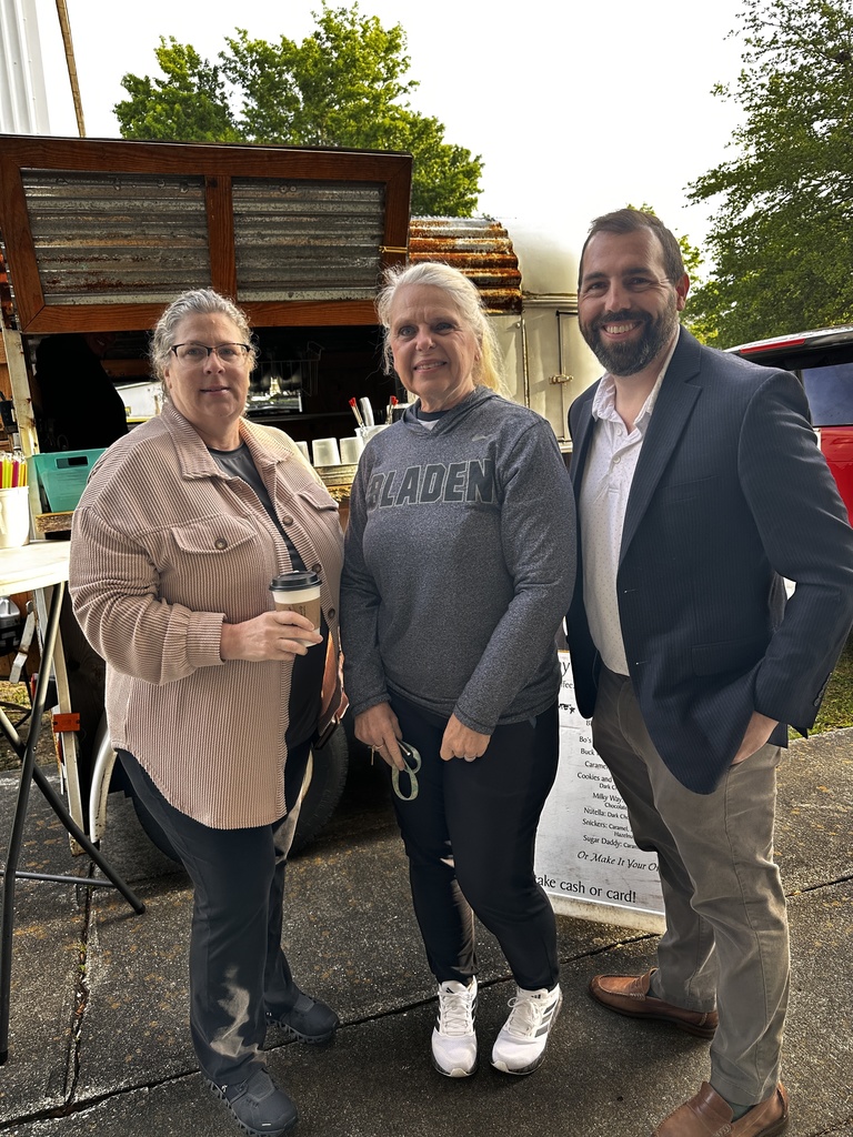Three people — two women and a man in a navy blazer — stand smiling in front of the Barefoot Brew coffee trailer. The woman in the center wears a gray "Bladen" sweatshirt. A menu board is visible in the background.