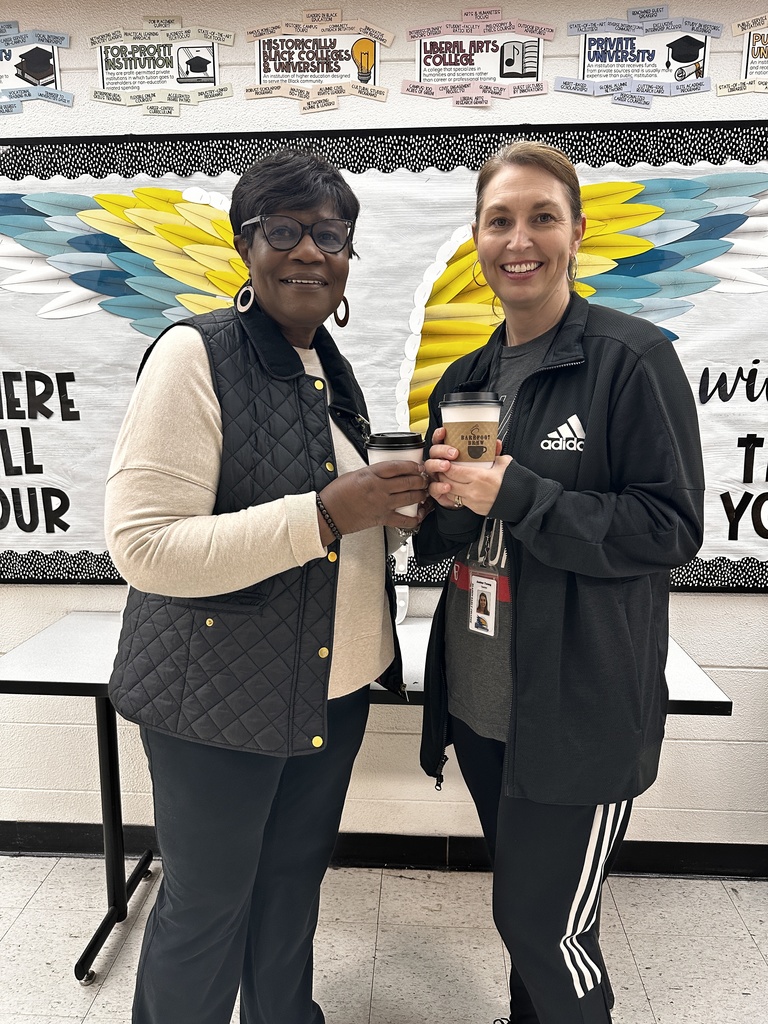 Two women stand together smiling and clinking their Barefoot Brew coffee cups in a school hallway. Behind them is a colorful bulletin board featuring different types of colleges and universities, including Historically Black Colleges & Universities, Liberal Arts College, and Private University, decorated with large painted wings.