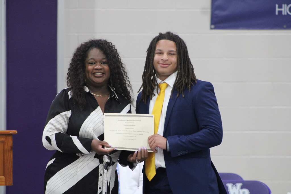 A student in a blue suit and yellow tie smiles while holding a scholarship certificate alongside a school administrator during West Bladen High School’s Scholastic Awards and Scholarship Night.