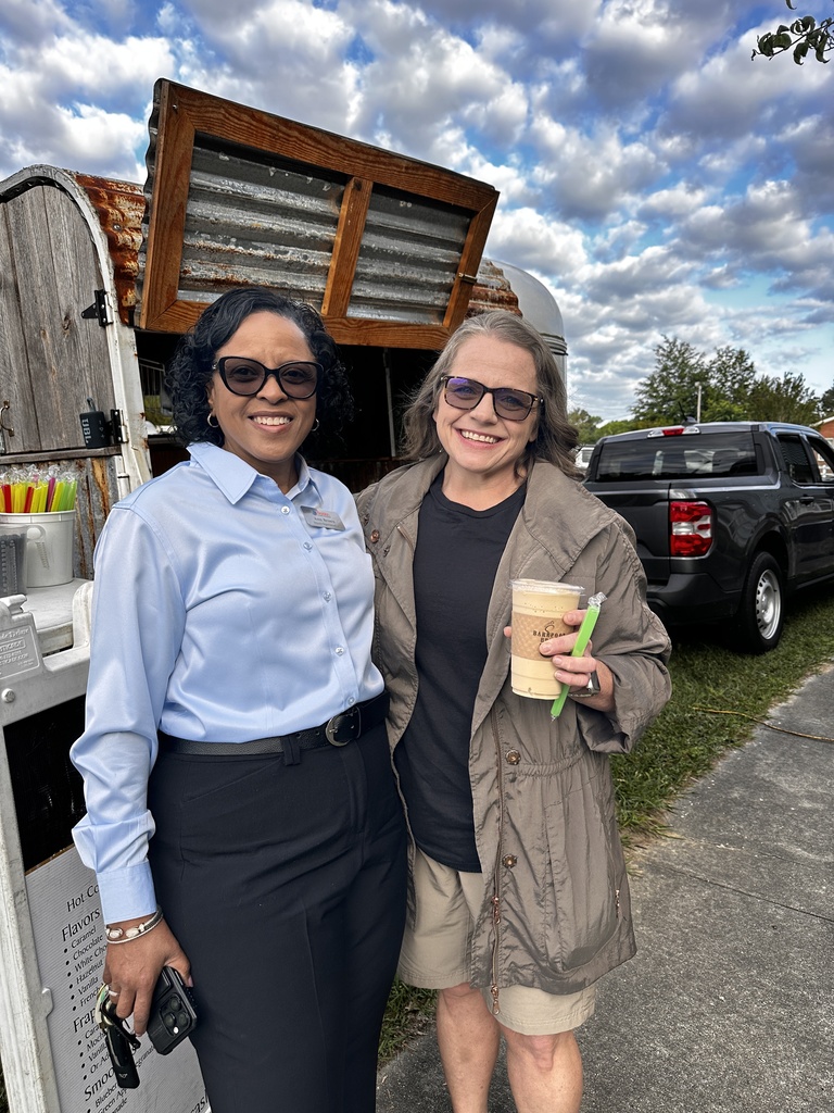 Two women stand smiling in front of a vintage coffee trailer. One wears a light blue button-down shirt; the other wears a tan jacket and holds an iced coffee drink.