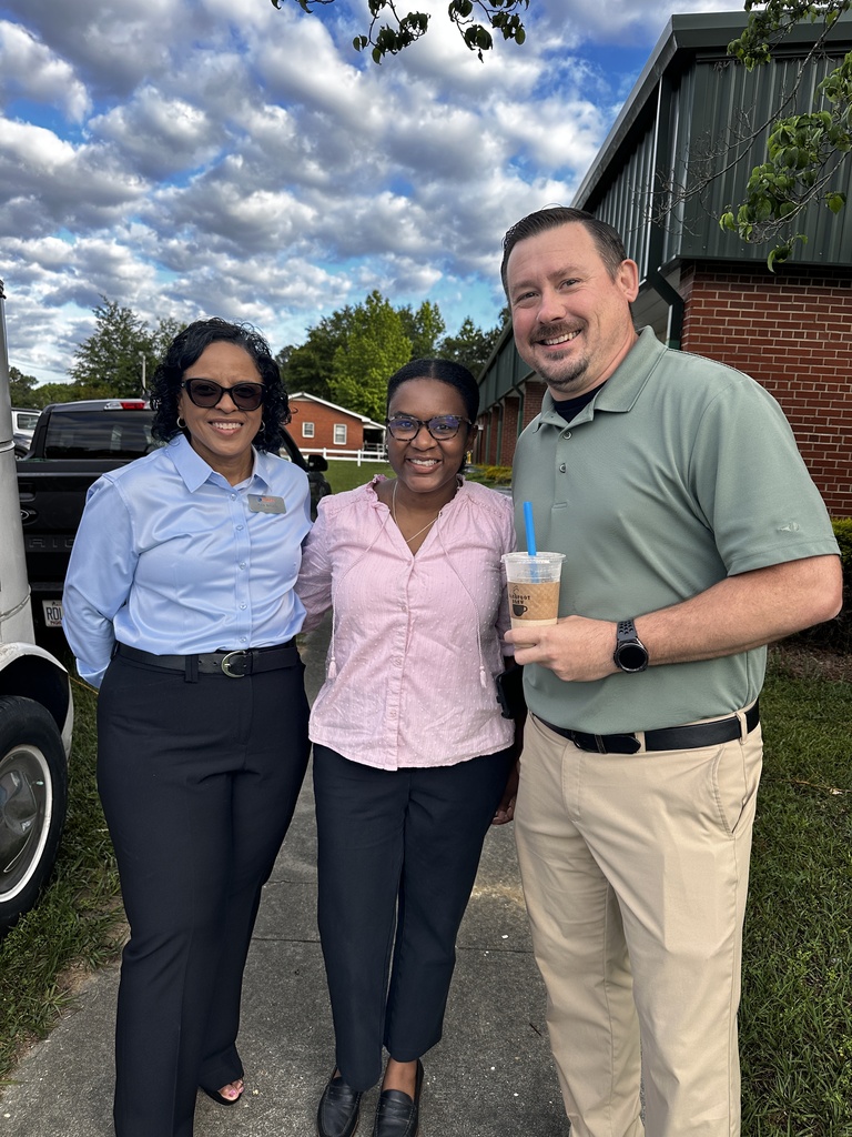 Three people stand together smiling outdoors beside a vintage truck. A woman in a light blue shirt, a woman in a pink blouse holding an iced drink, and a man in a sage green polo.