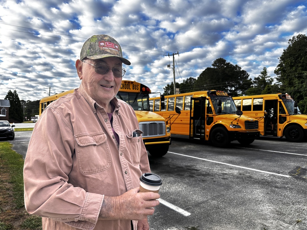 A man wearing a camo cap, glasses, and a pink button-down shirt holds a coffee cup and smiles outdoors, with a row of yellow school buses parked behind him.