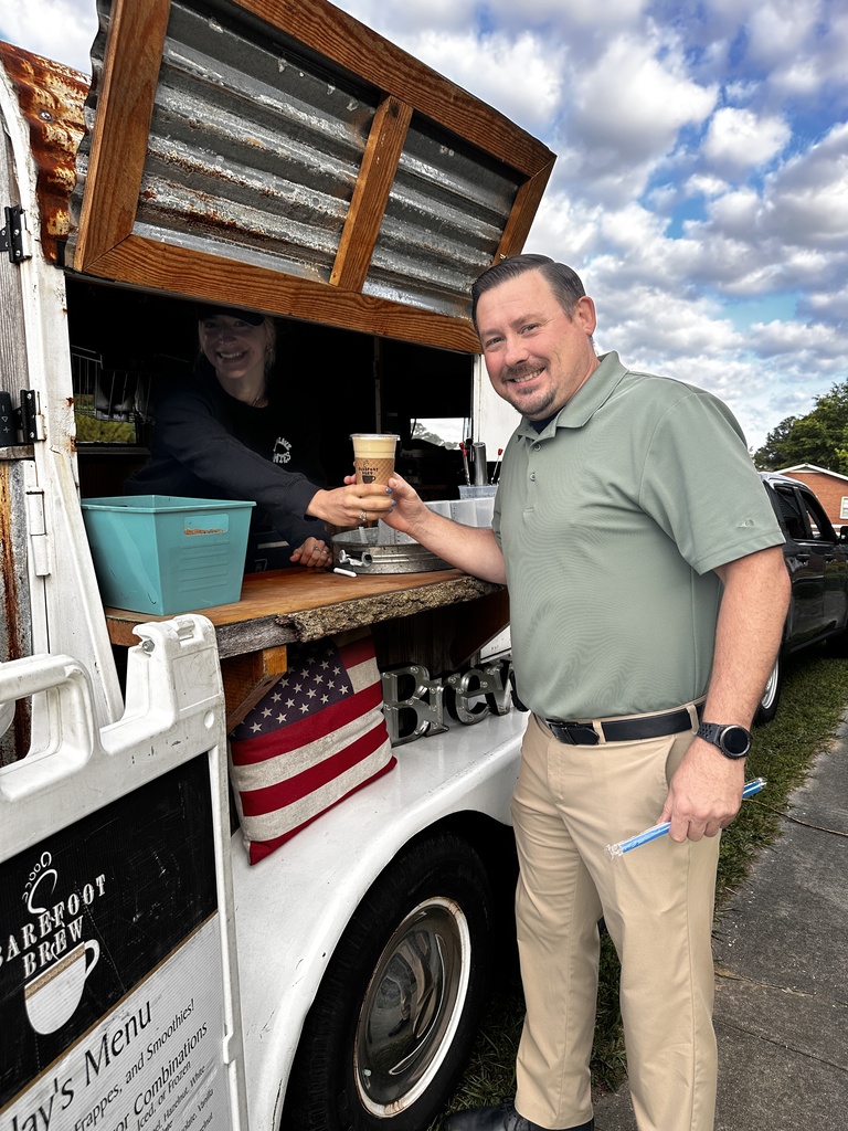 A man in a sage green polo shirt smiles as a barista hands him an iced coffee through the window of a Barefoot Brew vintage coffee trailer.