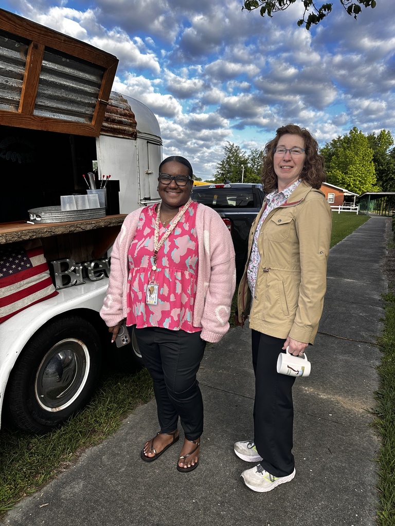 Two women stand smiling beside a vintage coffee trailer. One wears a pink floral top with a cardigan and a lanyard; the other wears a tan jacket and holds a white coffee mug.