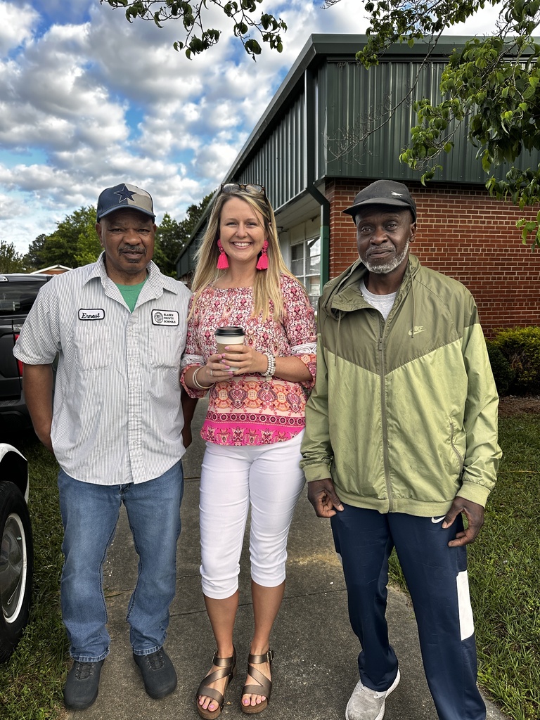 Three people stand smiling outdoors in front of a brick building. A woman in a pink floral top holds a coffee cup and stands between two men, one wearing a Bladen County Schools uniform shirt.