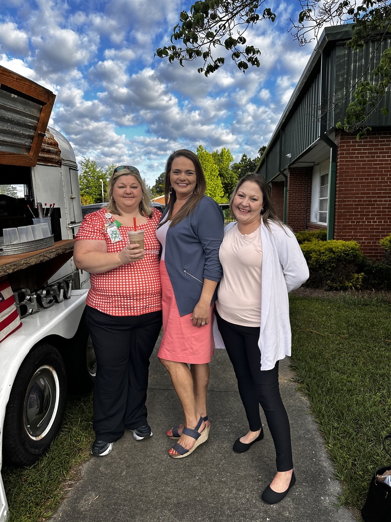 Three women pose together smiling beside a vintage coffee trailer. One holds an iced drink. They wear a red gingham top, a blue blazer with coral skirt, and a white cardigan.