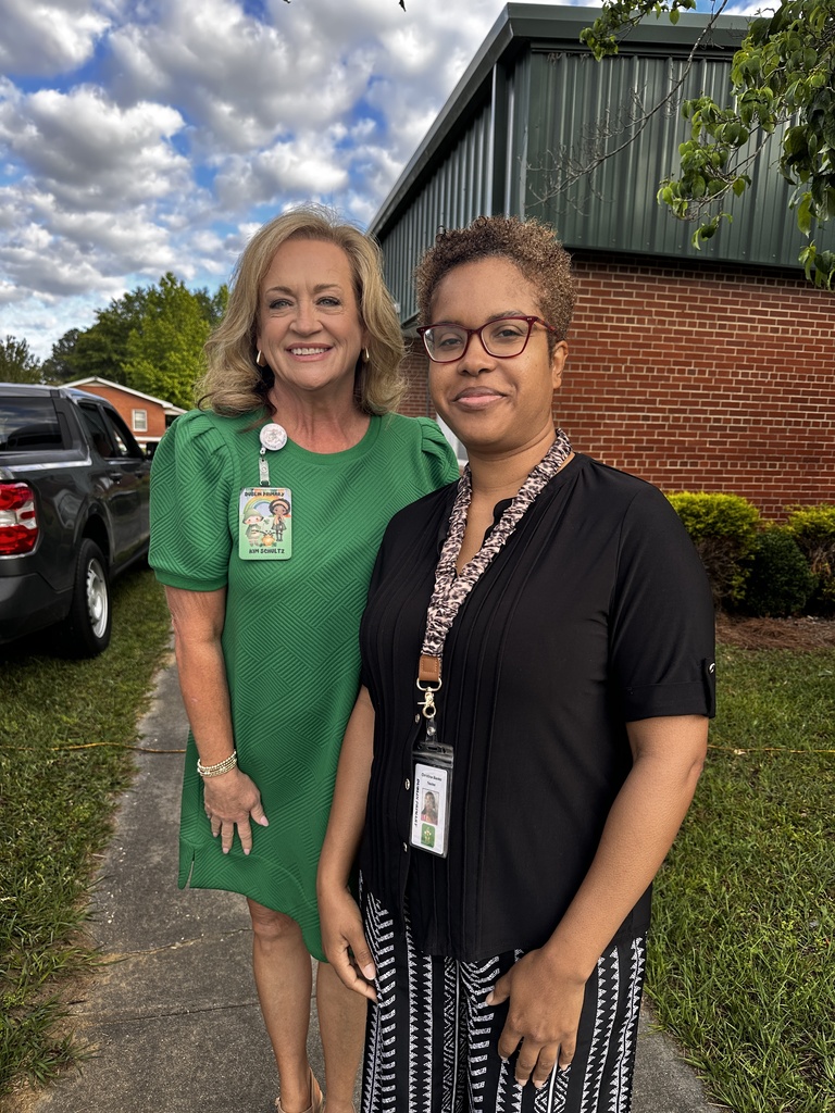 Two women stand smiling together outdoors in front of a brick building. One wears a green dress with a name badge; the other wears a black top with printed pants and a lanyard.