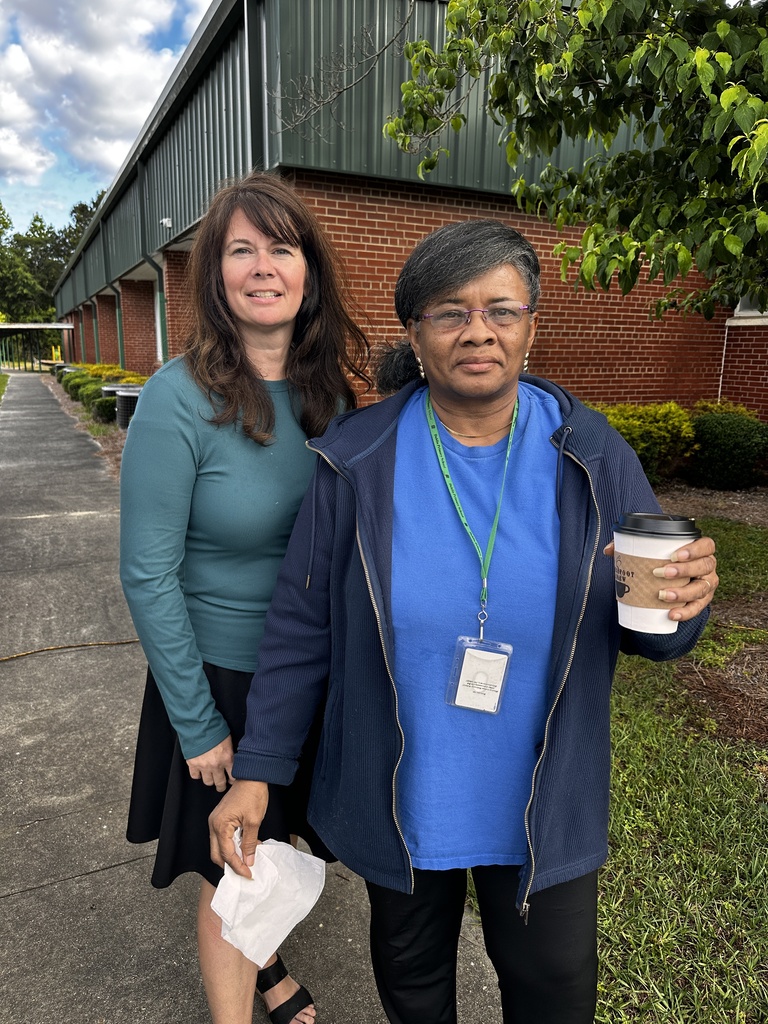 Two women stand together smiling outdoors in front of a brick building. One wears a teal long-sleeve top; the other wears a blue hoodie and holds a Barefoot Brew coffee cup.