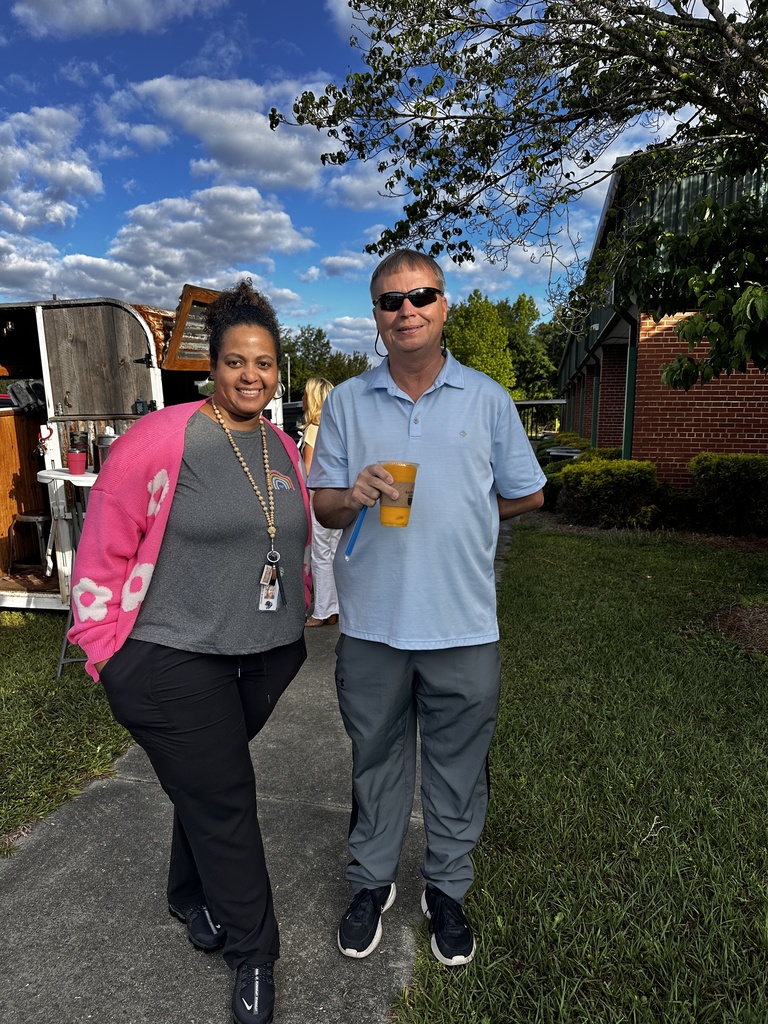 A woman in a pink floral cardigan and a man in a light blue polo shirt holding an orange iced drink stand together smiling outdoors near a coffee trailer.