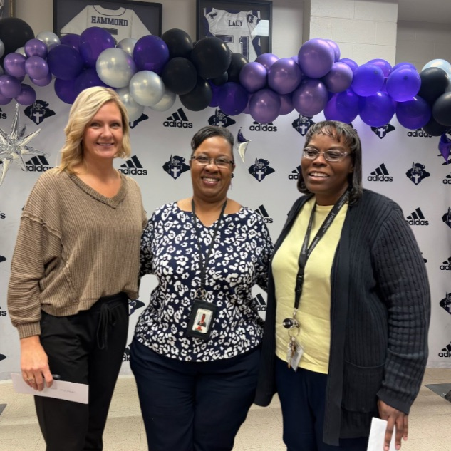 three women stand smiling in front of a school branded backdrop