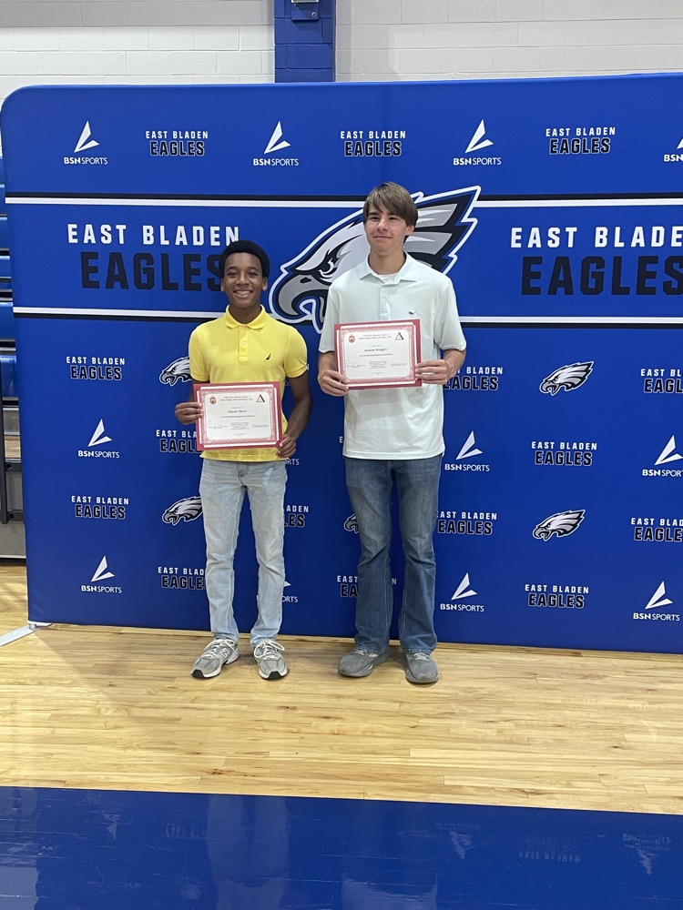 A student in a yellow shirt with a student in a white polo standing in front of the East Bladen backdrop posing with certificate certificates