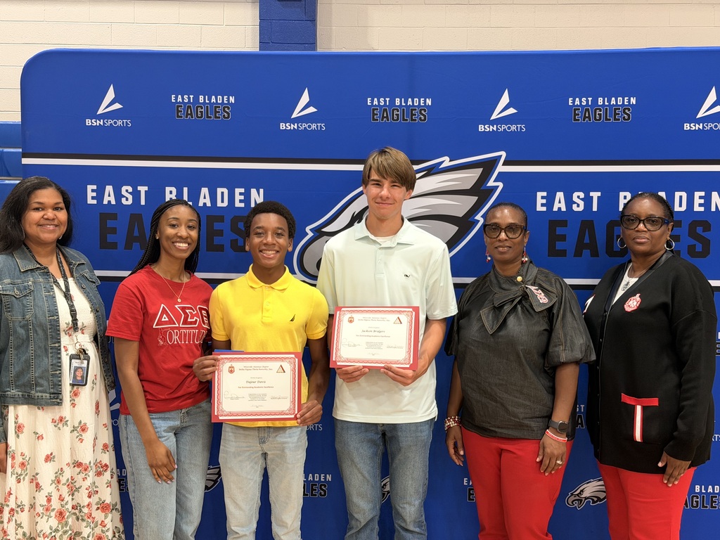 A student in a yellow shirt with a student in a white polo standing in front of the East Bladen backdrop posing with certificate certificates.  4 women in red also stand with the students.