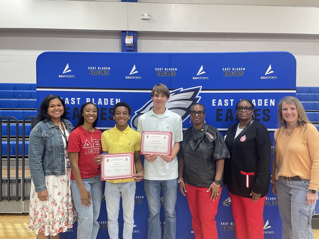 A student in a yellow shirt with a student in a white polo standing in front of the East Bladen backdrop posing with certificate certificates. A woman in an orange shirt and 4 women in red also stand with the students.