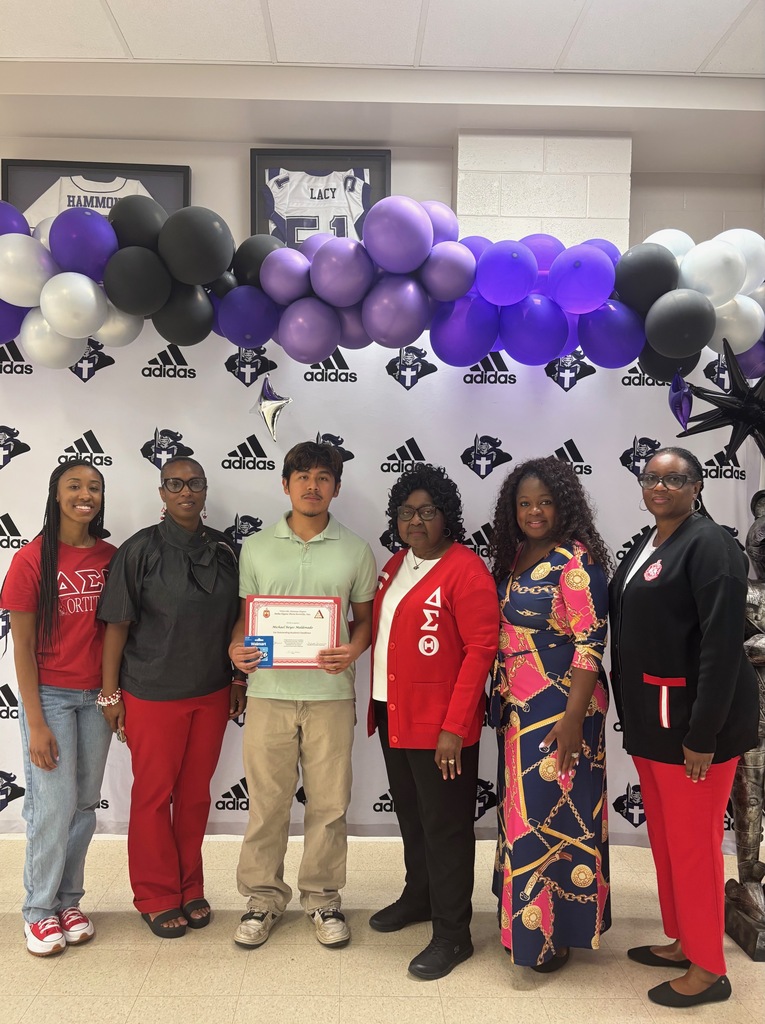 A group of seven people posing for a photo in front of a white backdrop featuring the West Bladen High School Knights and Adidas logos. In the center, a young man in a light green polo shirt holds a merit certificate and a gift card. He is flanked by five women, several of whom are wearing red and white attire representing Delta Sigma Theta Sorority, Inc., including one woman inA group of seven people posing for a photo in front of a white backdrop featuring the West Bladen High School Knights and Adidas logos. In the center, a young man in a light green polo shirt holds a merit certificate and a gift card. He is flanked by five women, several of whom are wearing red and white attire representing Delta Sigma Theta Sorority, Inc., including one woman in a red blazer with the Greek letters "ΔΣΘ a red blazer with the Greek letters "ΔΣΘ