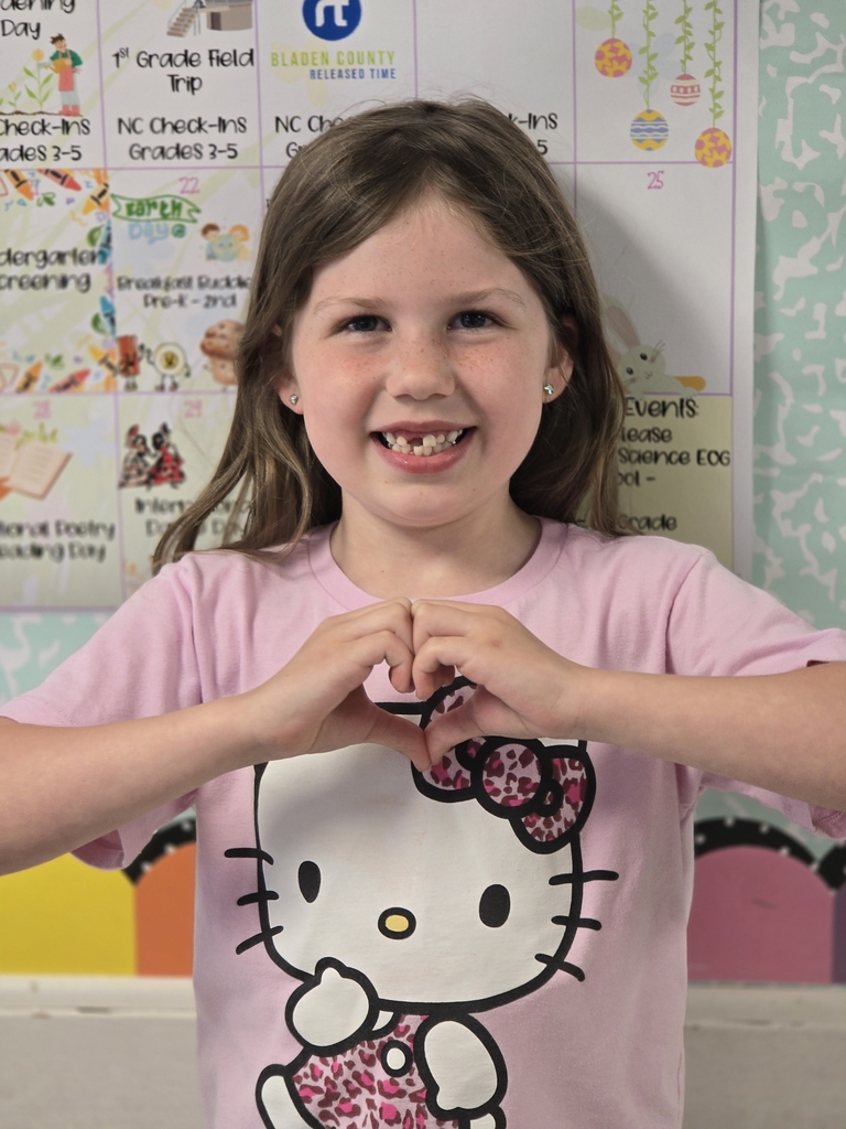 A smiling young girl wearing a pink Hello Kitty shirt poses in front of a classroom calendar while making a heart shape with her hands. She has long brown hair, freckles, and stud earrings, and colorful classroom decorations are visible in the background.
